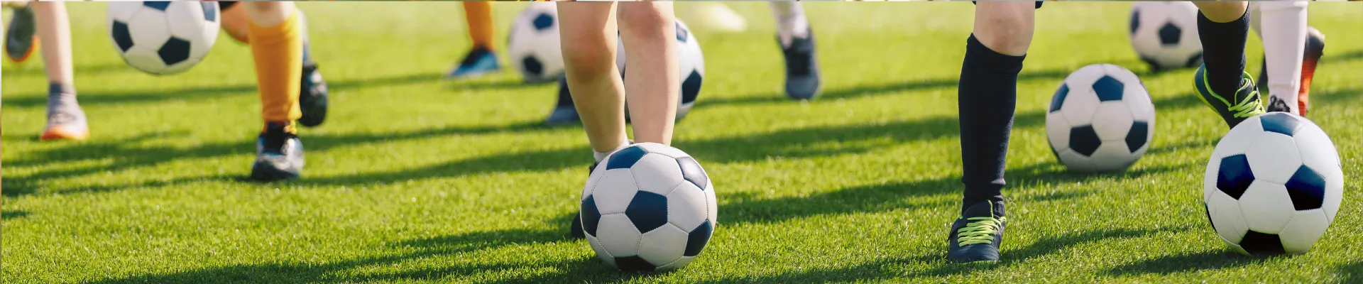 Children playing football on a grassy field with multiple soccer balls, focusing on their legs and feet.